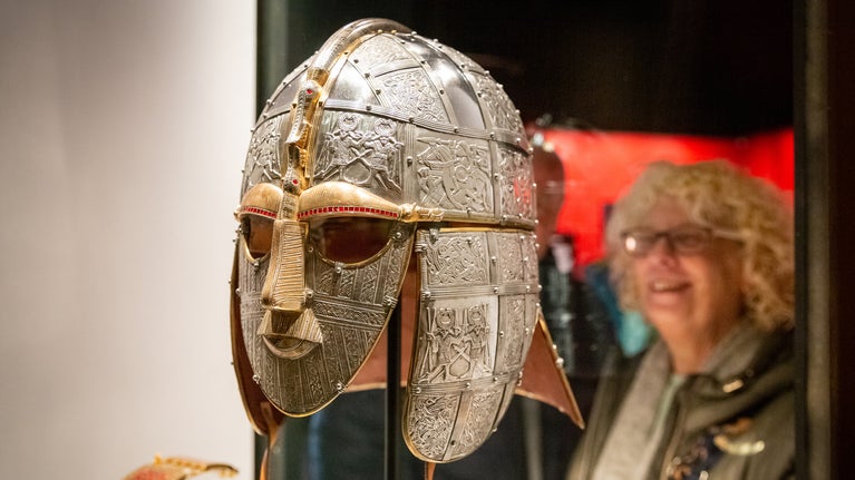 In the foreground is a metal helmet exquisitely worked into a mask of a man's face. Behind a lady is looking at the helmet, on display at the Exhibition Hall at Sutton Hoo, Suffolk.